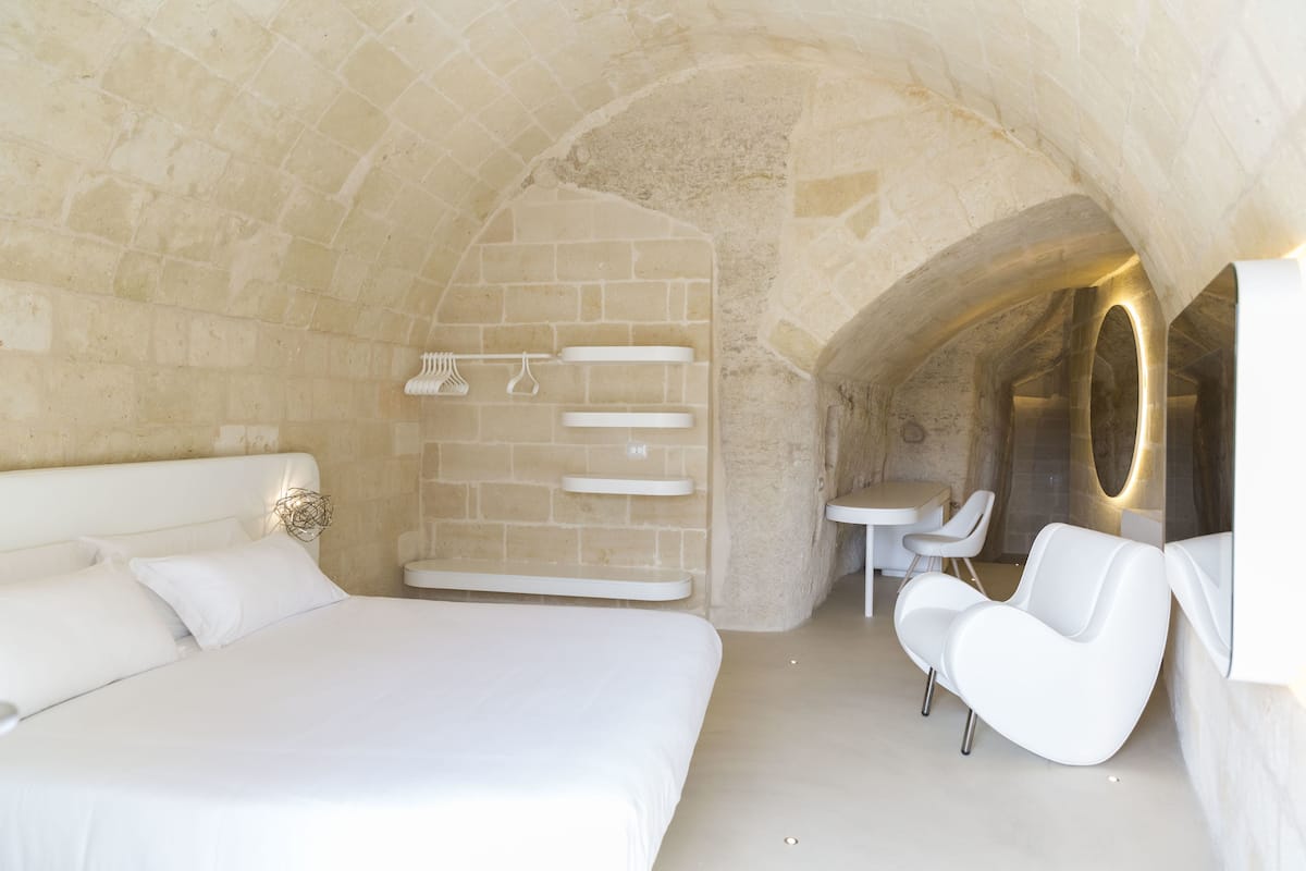 A modern bedroom with a stone arch, a large bed, a white chair, and a small table.