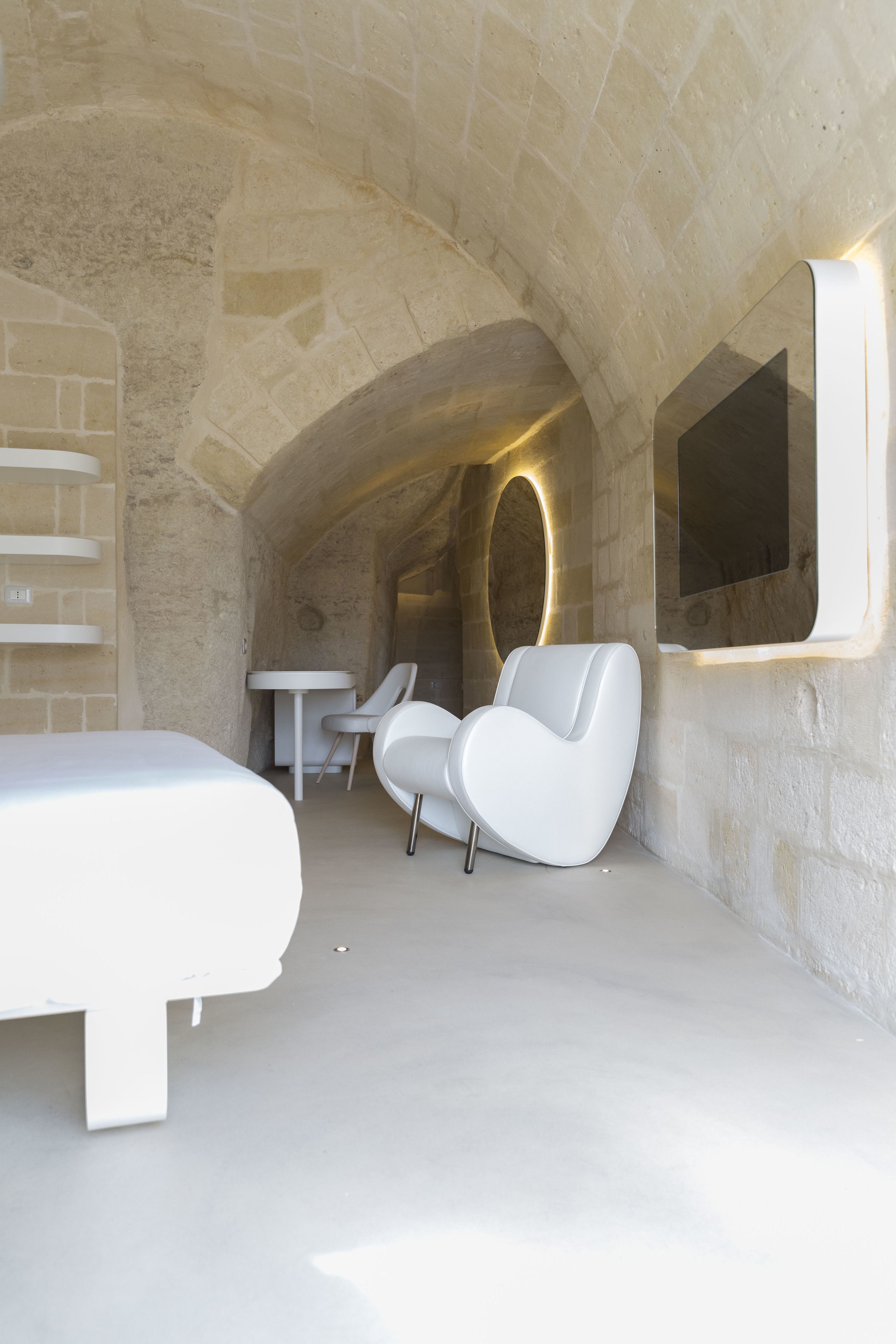 Une chambre à coucher moderne et minimaliste avec un plafond en forme d’arc en pierre, un lit, une chaise blanche et une petite table.