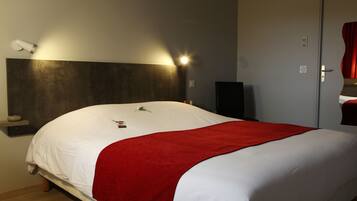 Una cama bien hecha con una colcha blanca y roja, una mesita de noche con una lámpara y un altavoz en el suelo.
