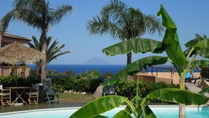 Zona de piscina con sillones reclinables, una estructura con techo de paja y palmeras.