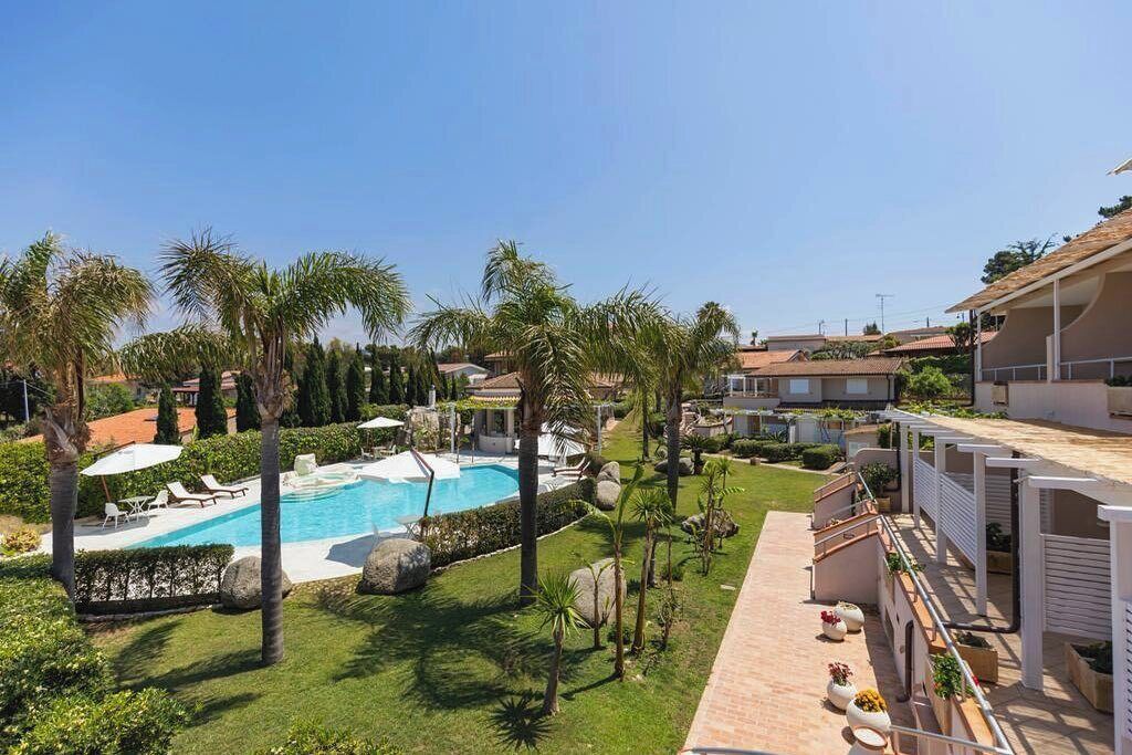 Zona de piscina con palmeras, cielo despejado y un edificio con tejado de baldosas.