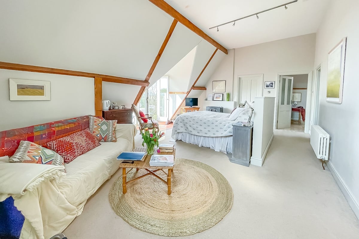 Ein geräumiges Zimmer mit hohen Decken, einem Bett, einem Sofa, einem Tisch mit einem Laptop und Blick ins Freie.