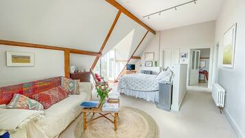 Una habitación amplia con techo alto, una cama, un sofá, una mesa con una computadora portátil y vistas al exterior.