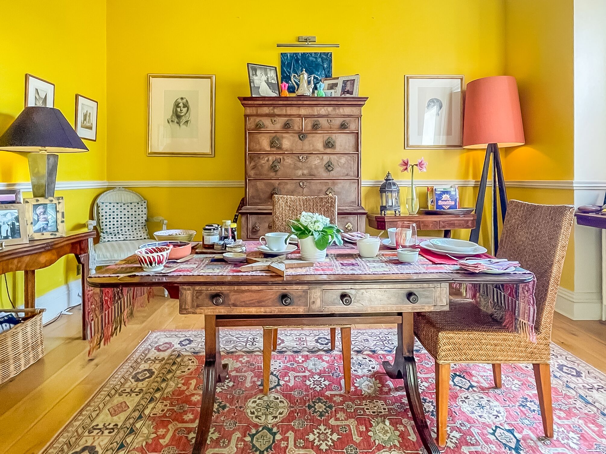 A dining area with a wooden table set for four, surrounded by chairs, a patterned rug, and a wooden chest of drawers.