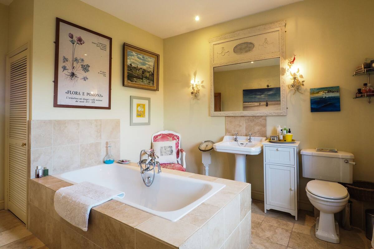 Ein Badezimmer mit einer großen Badewanne, einem Waschbecken und einer Toilette. An den Wänden hängen Bilder in Rahmen und über dem Waschbecken befindet sich ein Spiegel.