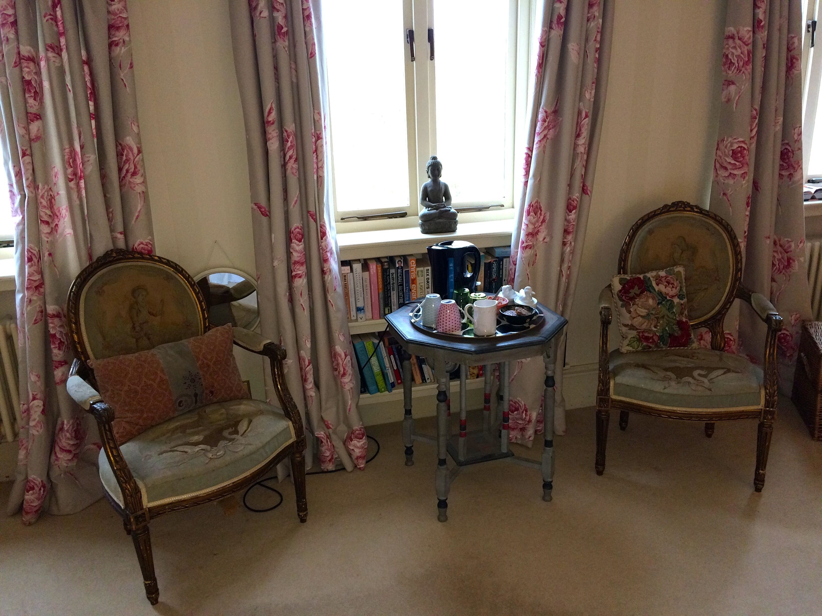 A room with two ornate chairs, a small table with a teapot and cups, and a bookshelf filled with books.