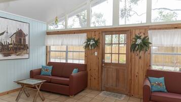 A cozy room with a wooden door, two red sofas with blue cushions, a glass coffee table, and a large window with white curtains.