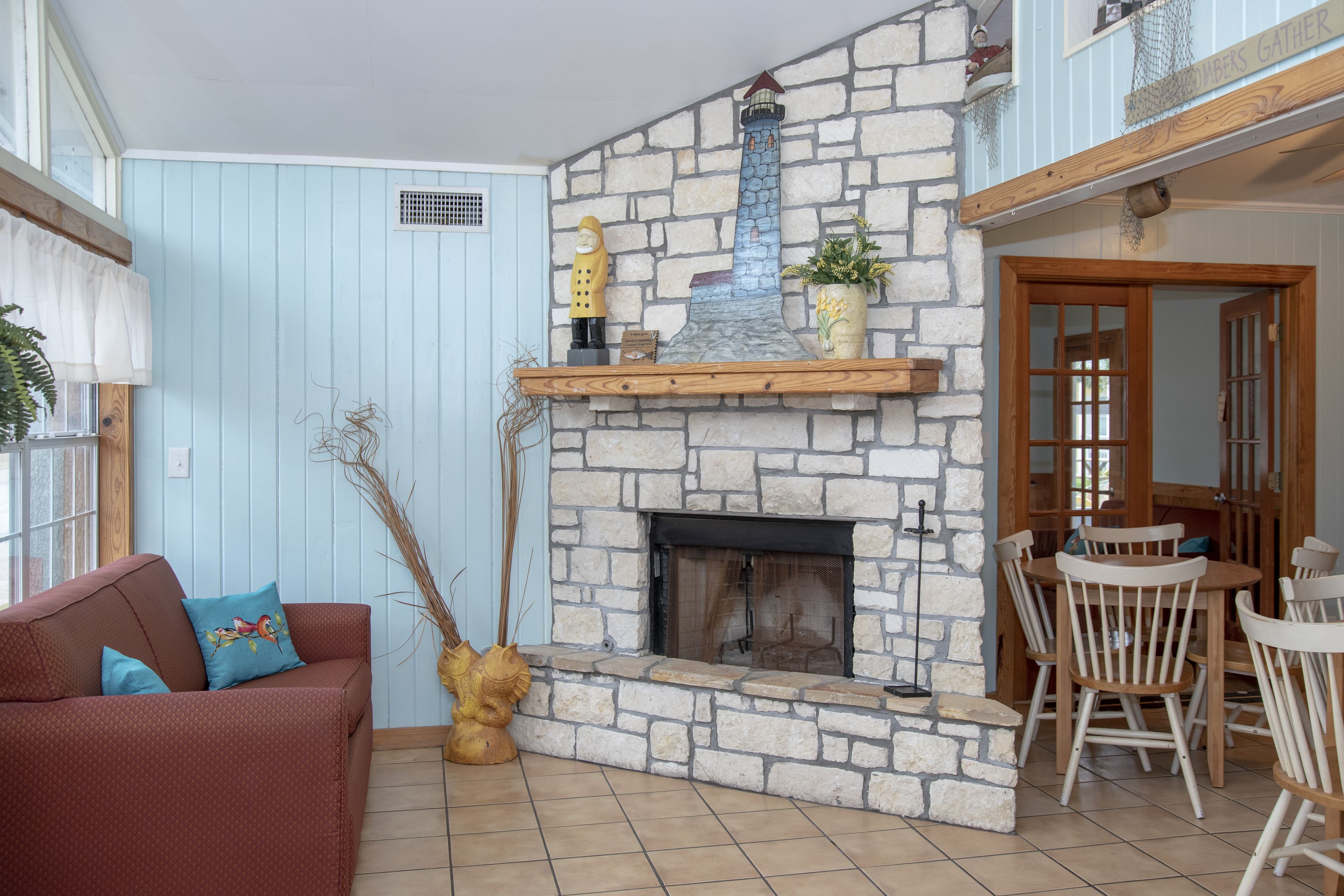 A cozy living room with a stone fireplace, a wooden dining table with chairs, a red sofa with a blue pillow, and a wooden floor.
