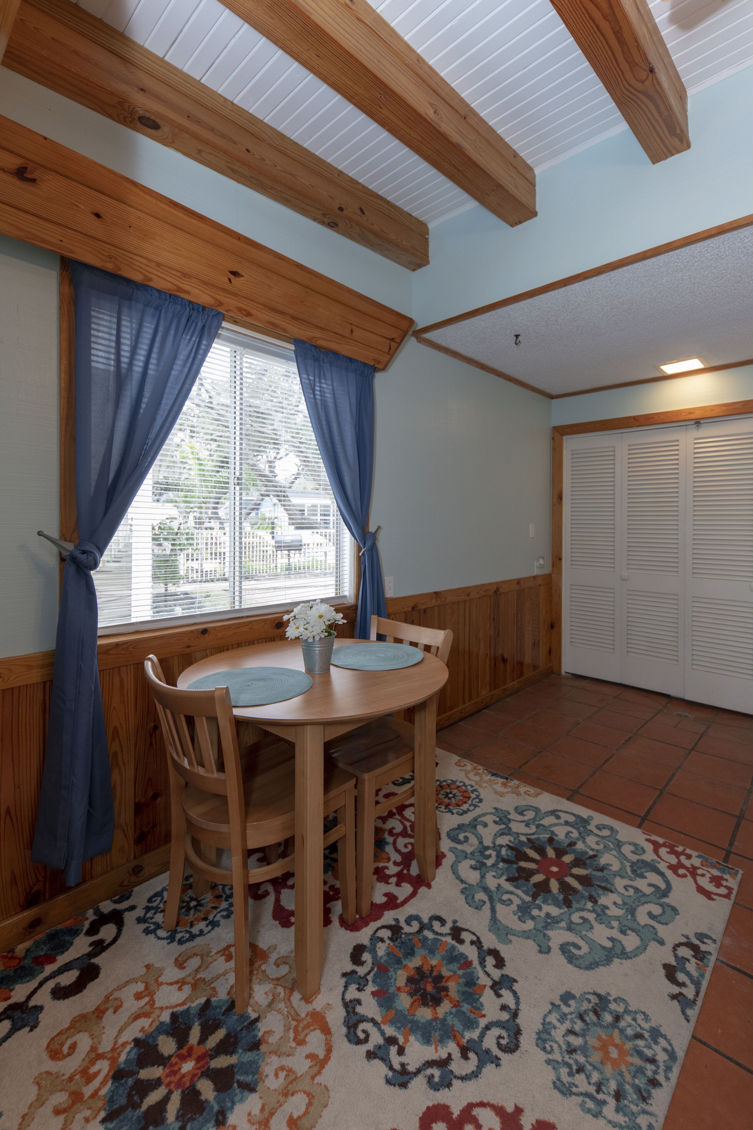 A room with a wooden dining table, two chairs, a vase with flowers, and a patterned rug.