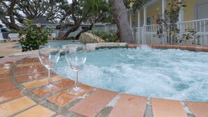 Een jacuzzi met twee glazen water, omringd door een betegeld terras en een geel gebouw met een witte veranda.