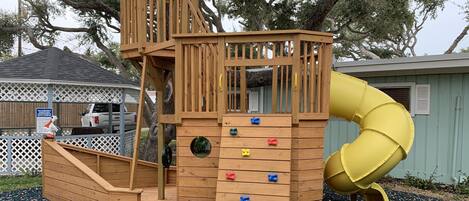 A wooden play structure with a slide and climbing wall, surrounded by trees and a gravel ground.