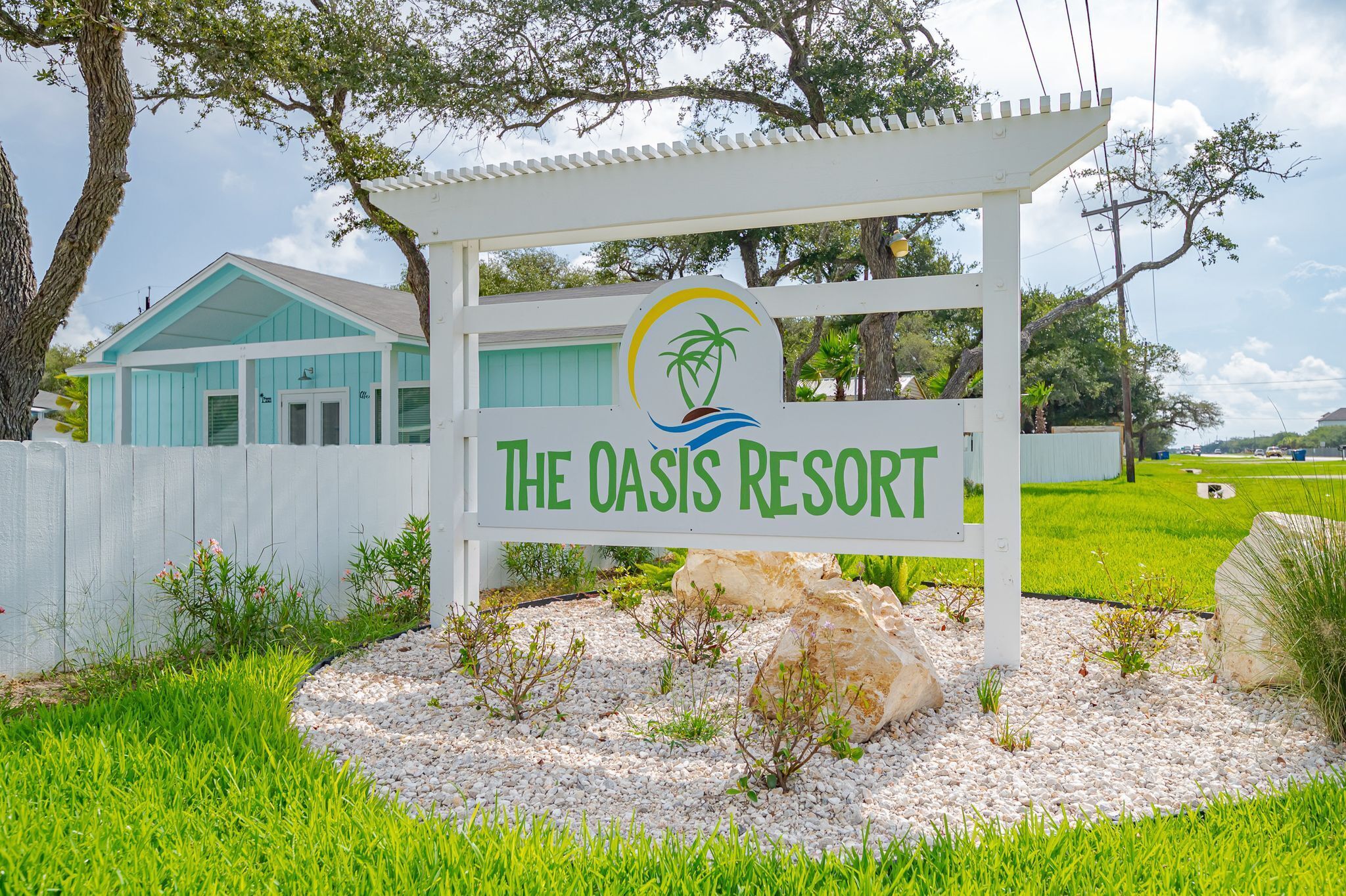 Señal del Resort Oasis, con logotipo, rodeada de follaje y una valla blanca.