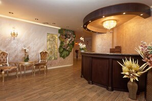 A reception area with a curved reception desk, two ornate chairs, a wall-mounted light fixture, and a large floral arrangement. - Elena Hotel - All Inclusive (Golden Sands)
