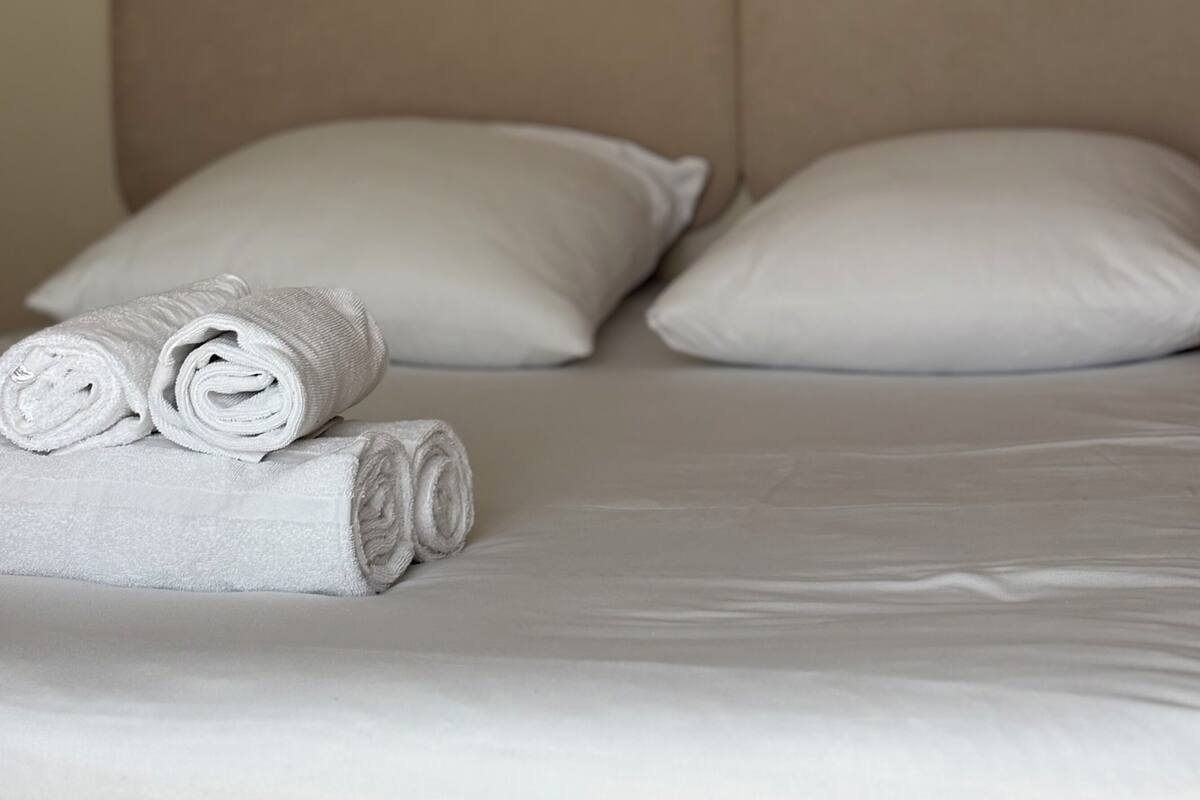 A neatly made bed with white linens and pillows, a wall socket above the bed, and a plain wall.