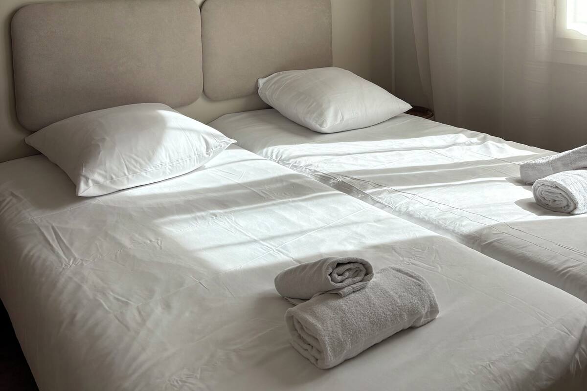 A hotel room with a bed, pillows, a wall-mounted light, and a window with curtains.