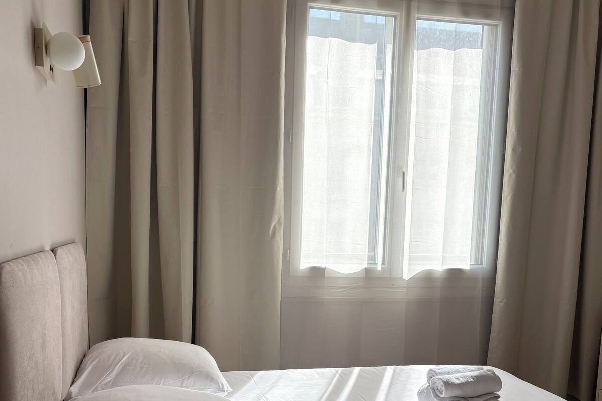 A neatly made bed with white linens, a round wooden side table, and a window with curtains.