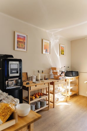 A kitchen with a coffee machine, a microwave, and a sink. There are framed pictures on the wall and a wooden table with fruit and dishes. - Hôtel Bloom (Caen)