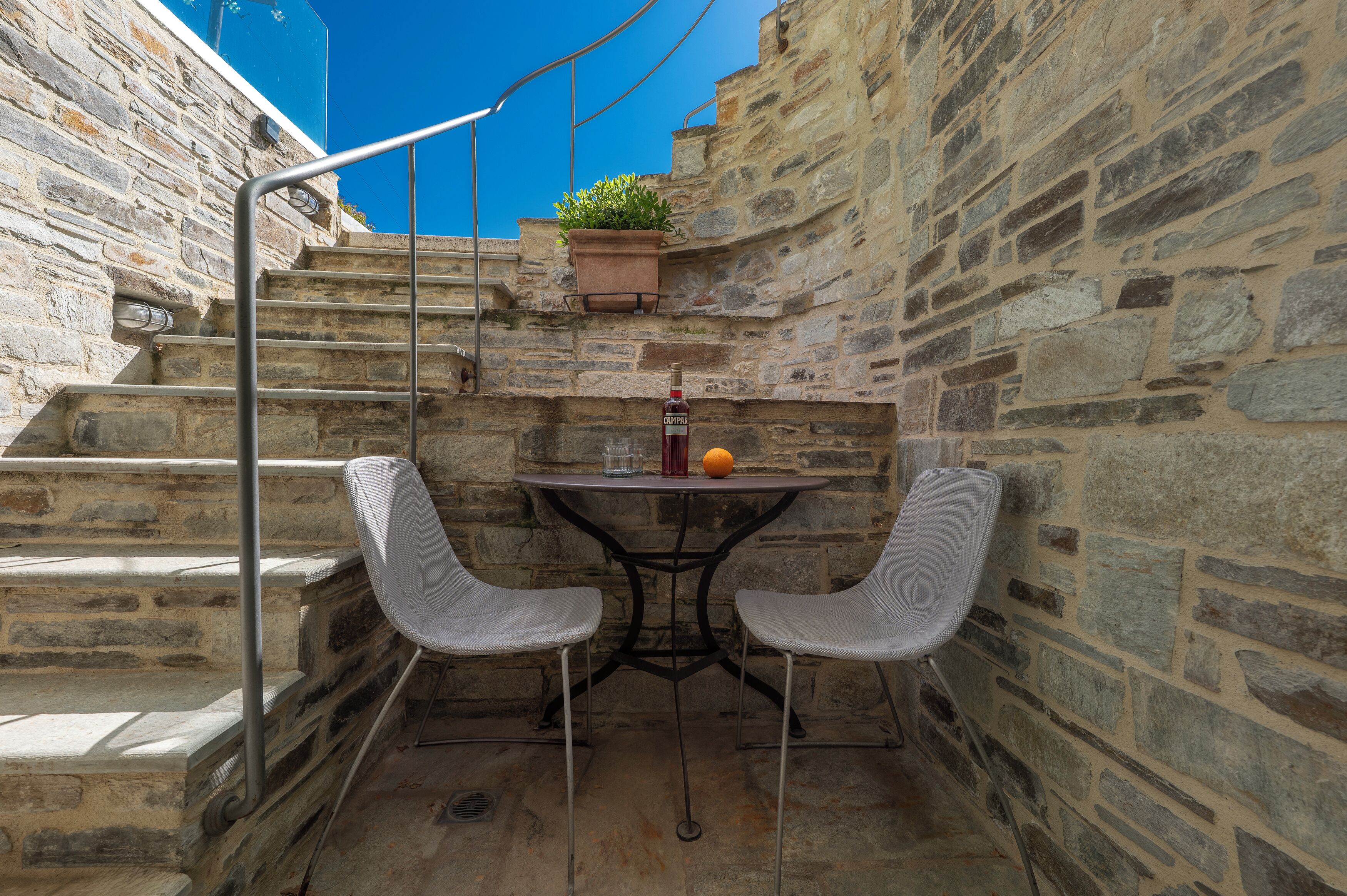 Un escalier en pierre mène à un petit espace extérieur aménagé avec deux chaises et une table, un pot de fleurs et une bouteille de vin.