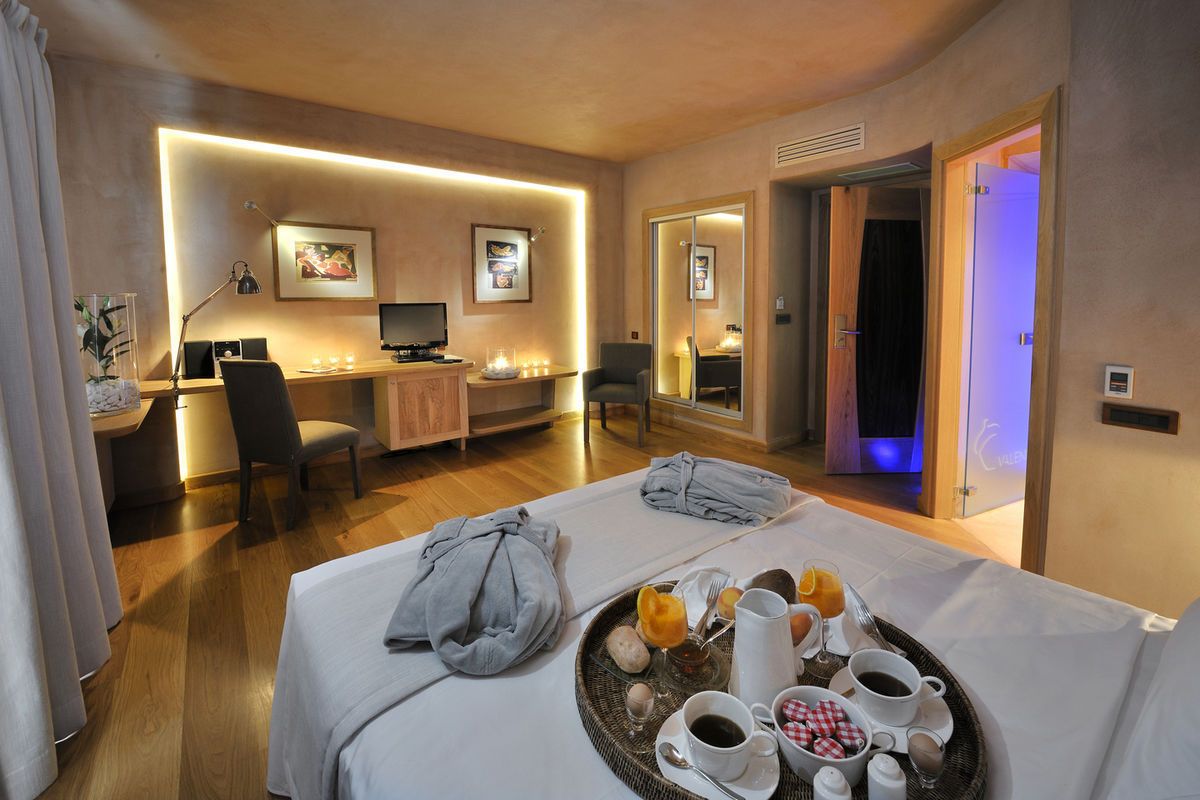 A hotel room with a dining table set for breakfast, a desk with a computer, and a bed with a tray of breakfast items.