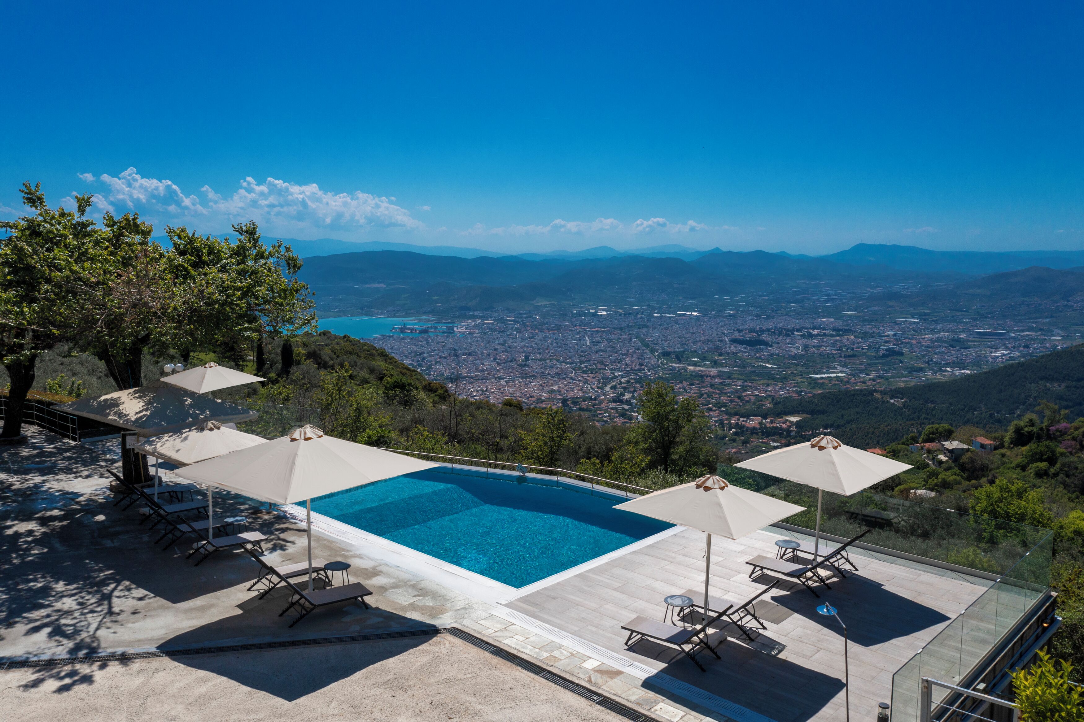 A rooftop infinity pool with lounge chairs and umbrellas, overlooking a cityscape and mountains.