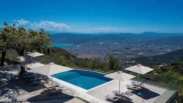 A rooftop infinity pool with lounge chairs and umbrellas, overlooking a cityscape and mountains.