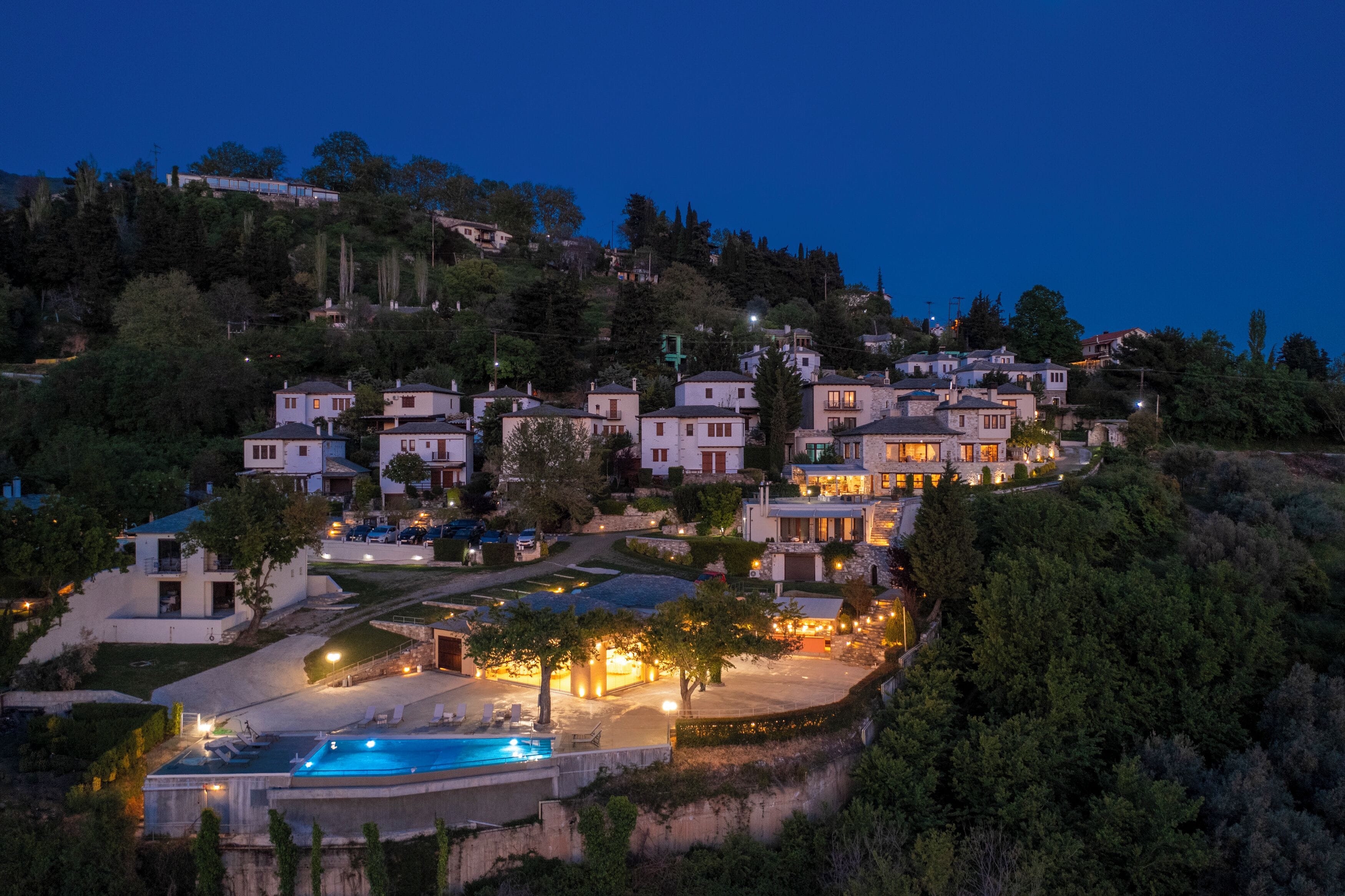 A hillside residential area with multiple houses, a swimming pool, and a well-lit driveway.