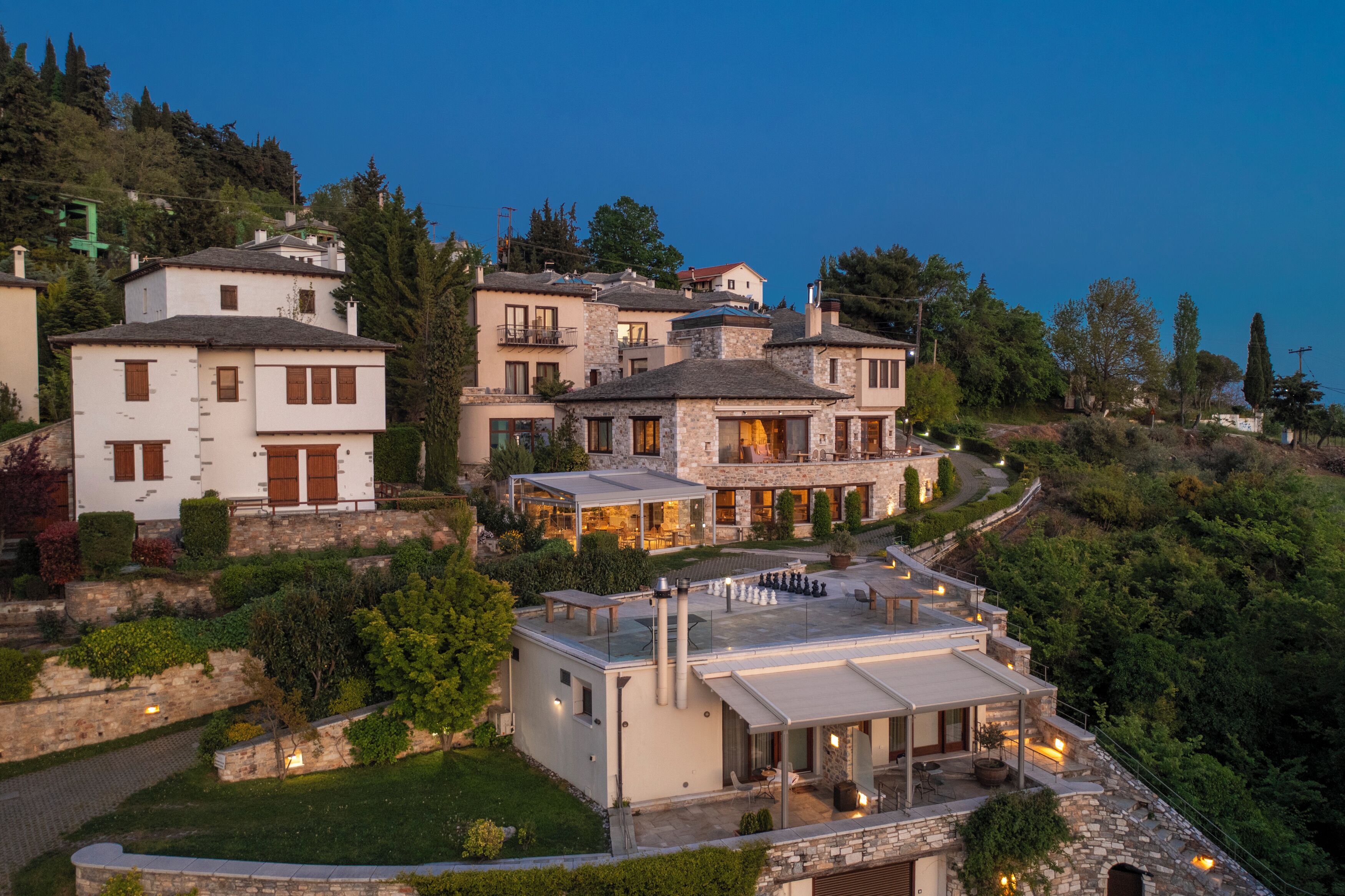 A hillside residential area with multiple houses, featuring stone and modern architecture, and a well-maintained garden.