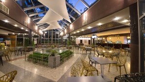 A spacious indoor area with a high ceiling, glass panels, and a central garden. There are multiple seating areas with tables and chairs, and a bar area in the background. - Mimosa Sunshine Hotel COOEE - All Inclusive (Golden Sands)
