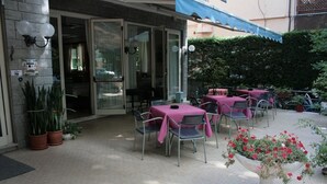An outdoor patio with tables and chairs, a potted plant, and a building entrance. - Hotel Ardesia (Rimini)