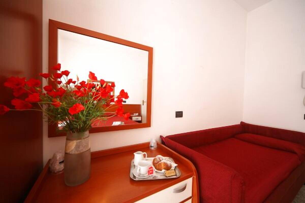 A hotel room with a wooden bedside table, a vase of red flowers, a tray with bread and a jug, and a red sofa.