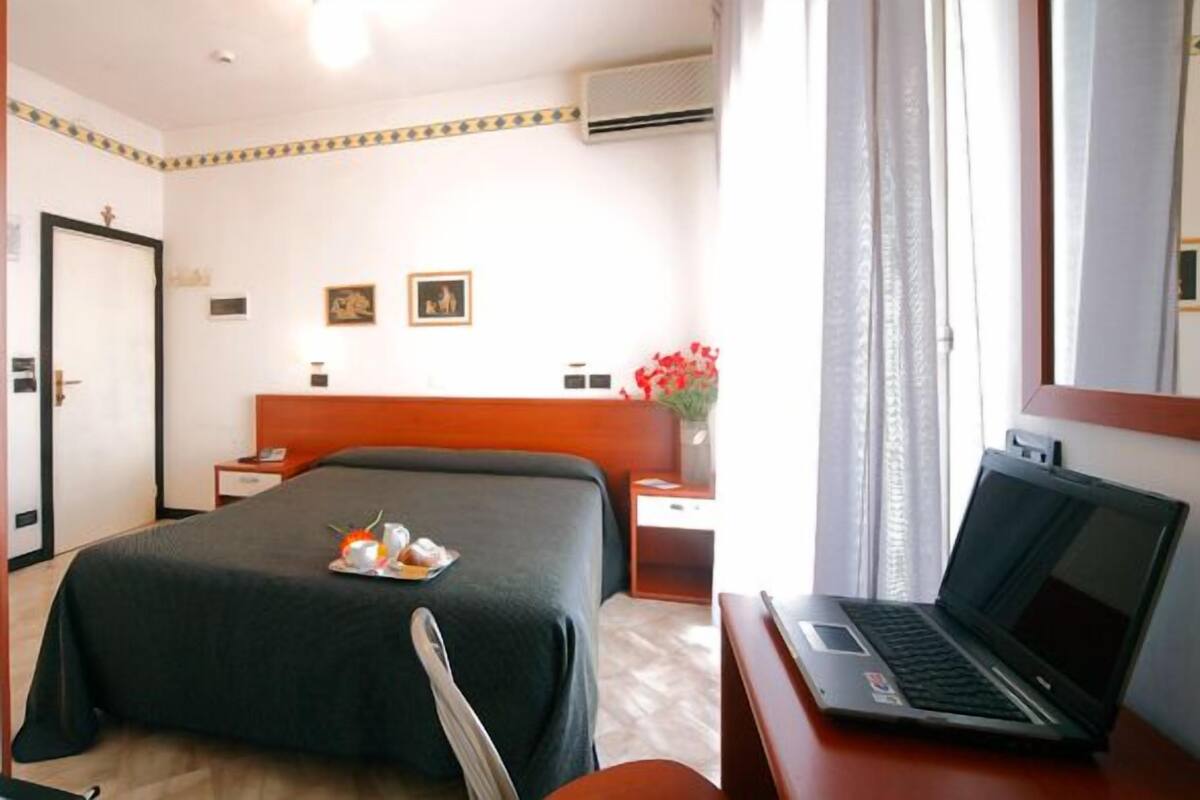 A hotel room with a bed, a desk with a laptop, a nightstand, a bedside table, a mirror, and a window with curtains.