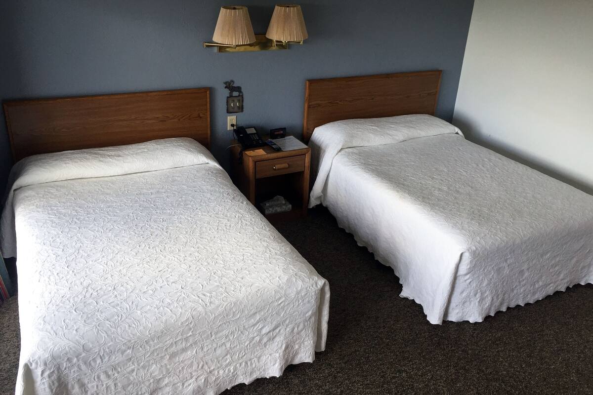 A hotel room with two single beds, a wooden headboard, a nightstand with a phone and lamp, and a wall-mounted light fixture.