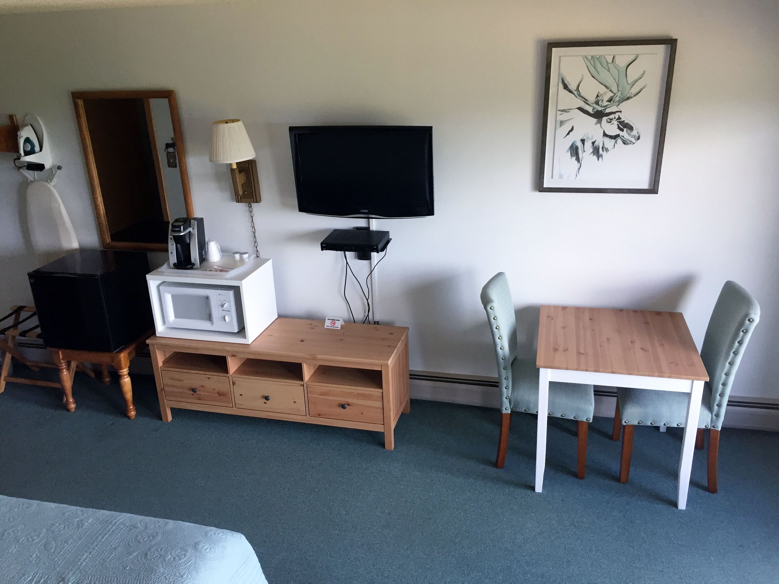 Una habitación de hotel con una cama, una mesa de madera, dos sillas, un televisor, un microondas, una lámpara, un espejo y un cuadro en la pared.