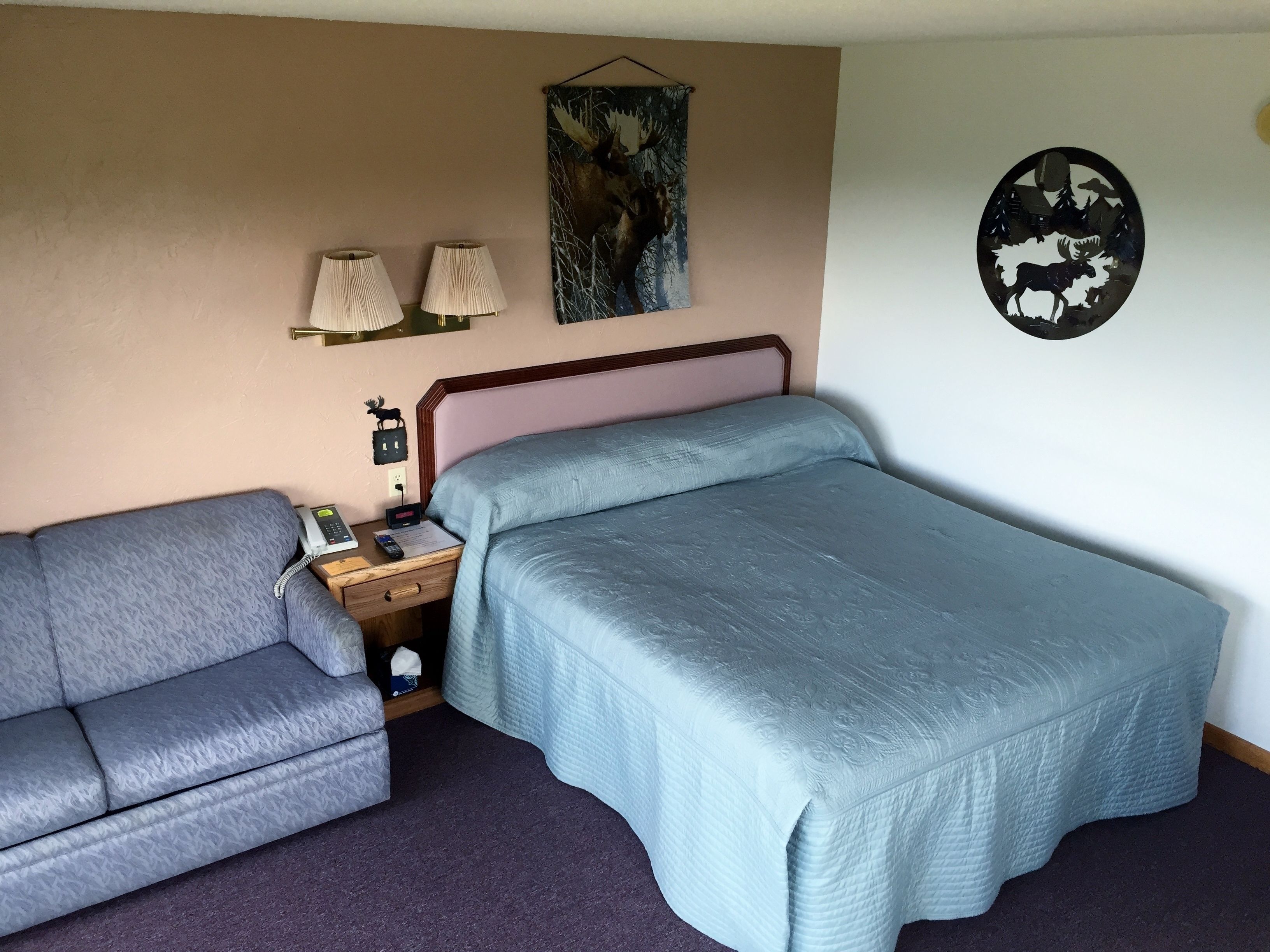 Habitación de hotel con cama, sofá, mesita de noche con lámpara, un adorno en la pared y un reloj de pared.