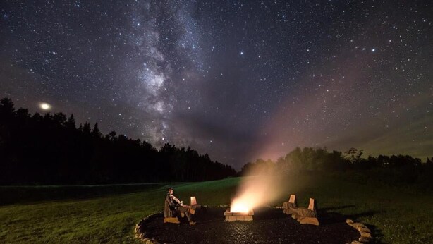 A night scene with a starry sky, a fire pit with stones, and a person sitting by the fire. - Kineo View Lodge (Greenville)