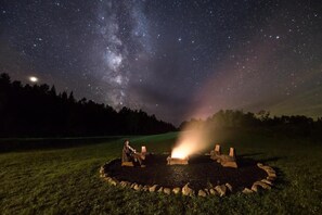 A night scene with a starry sky, a fire pit with stones, and a person sitting by the fire. - Kineo View Lodge (Greenville)