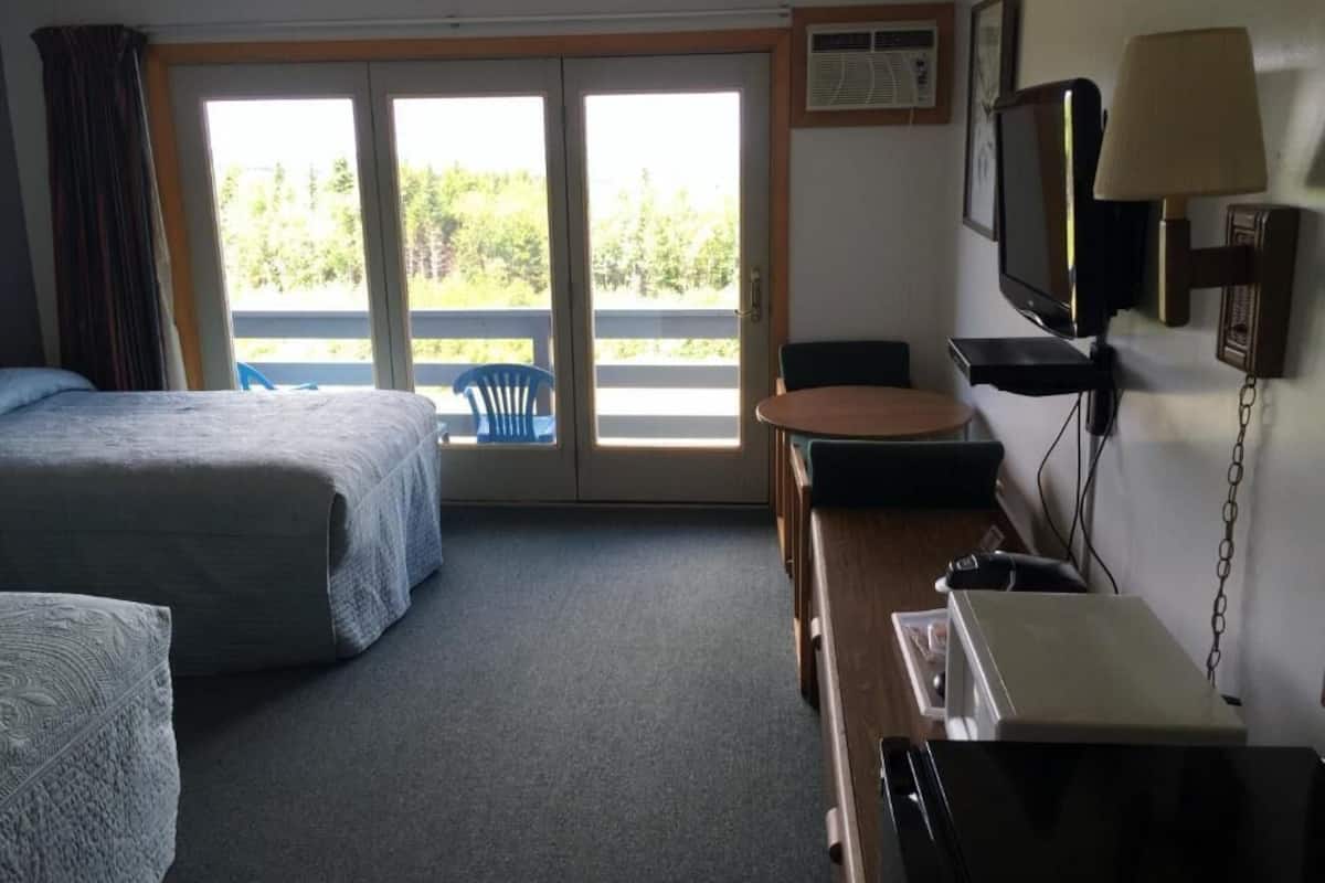A hotel room with two beds, a desk with a lamp, a television, and a view of the outdoors through a sliding door.