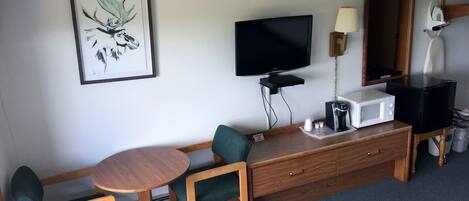 Habitación de hotel con televisión, cafetera, microondas, una mesita, dos sillas y una obra de arte enmarcada en la pared.
