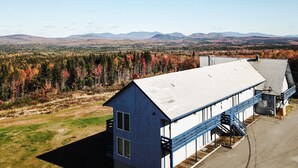 A large building with a flat roof and a blue exterior, surrounded by a forest with autumn foliage. - Kineo View Lodge (Greenville)