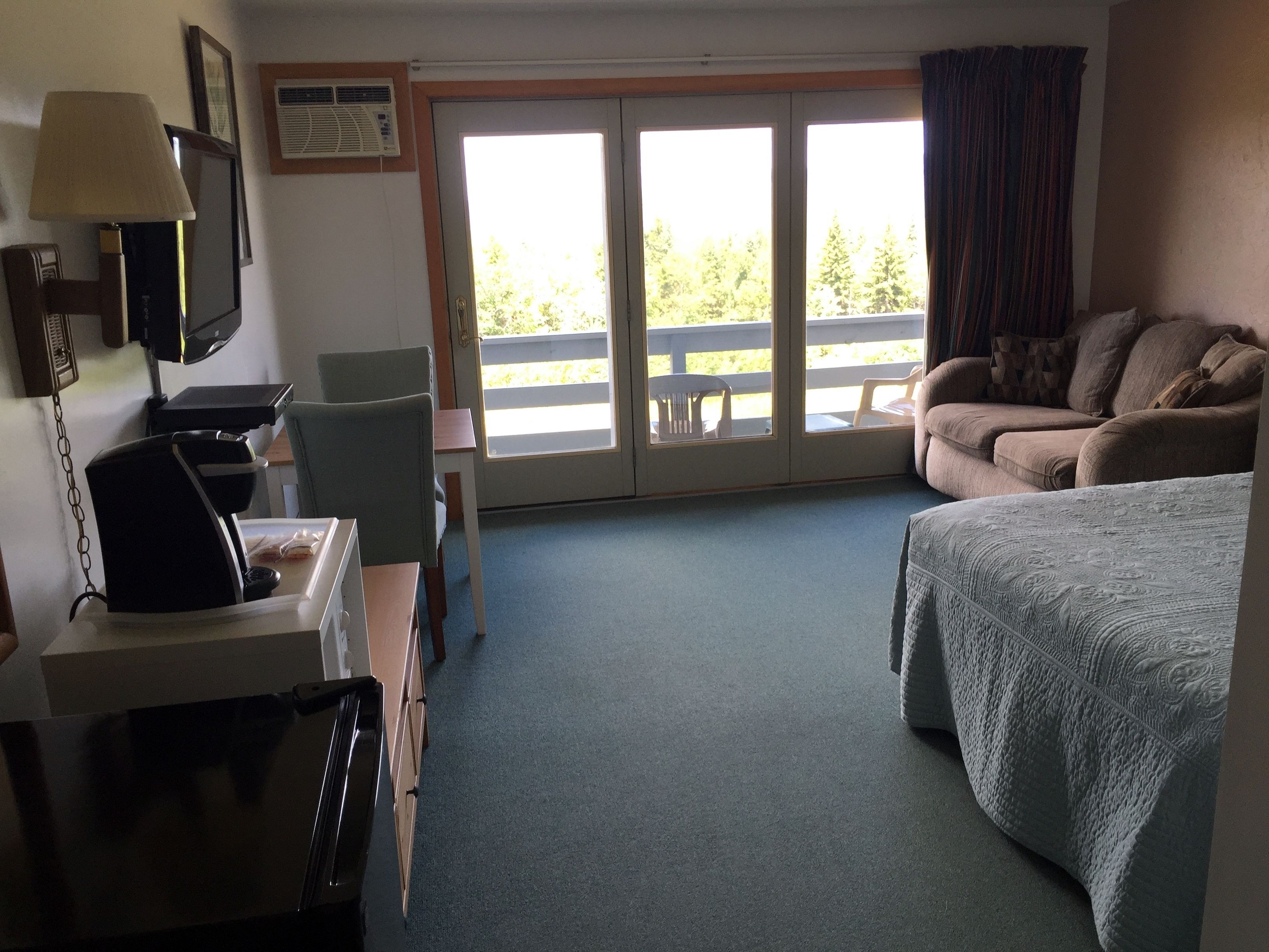 A hotel room with a bed, a desk with a lamp, a chair, a sofa, and a view of a balcony with outdoor furniture.