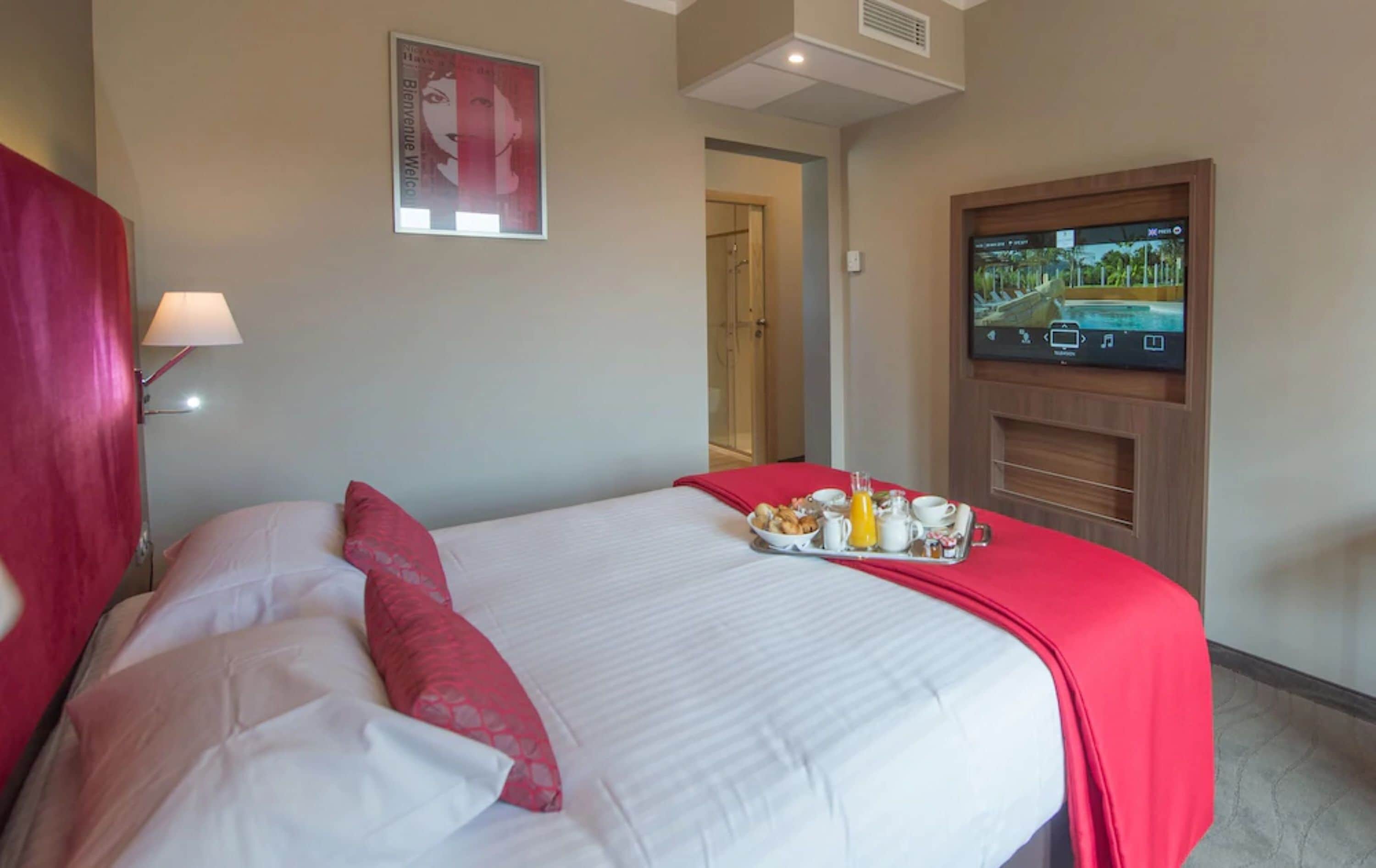A hotel room with a bed, a TV mounted on the wall, and a breakfast tray on the bed.