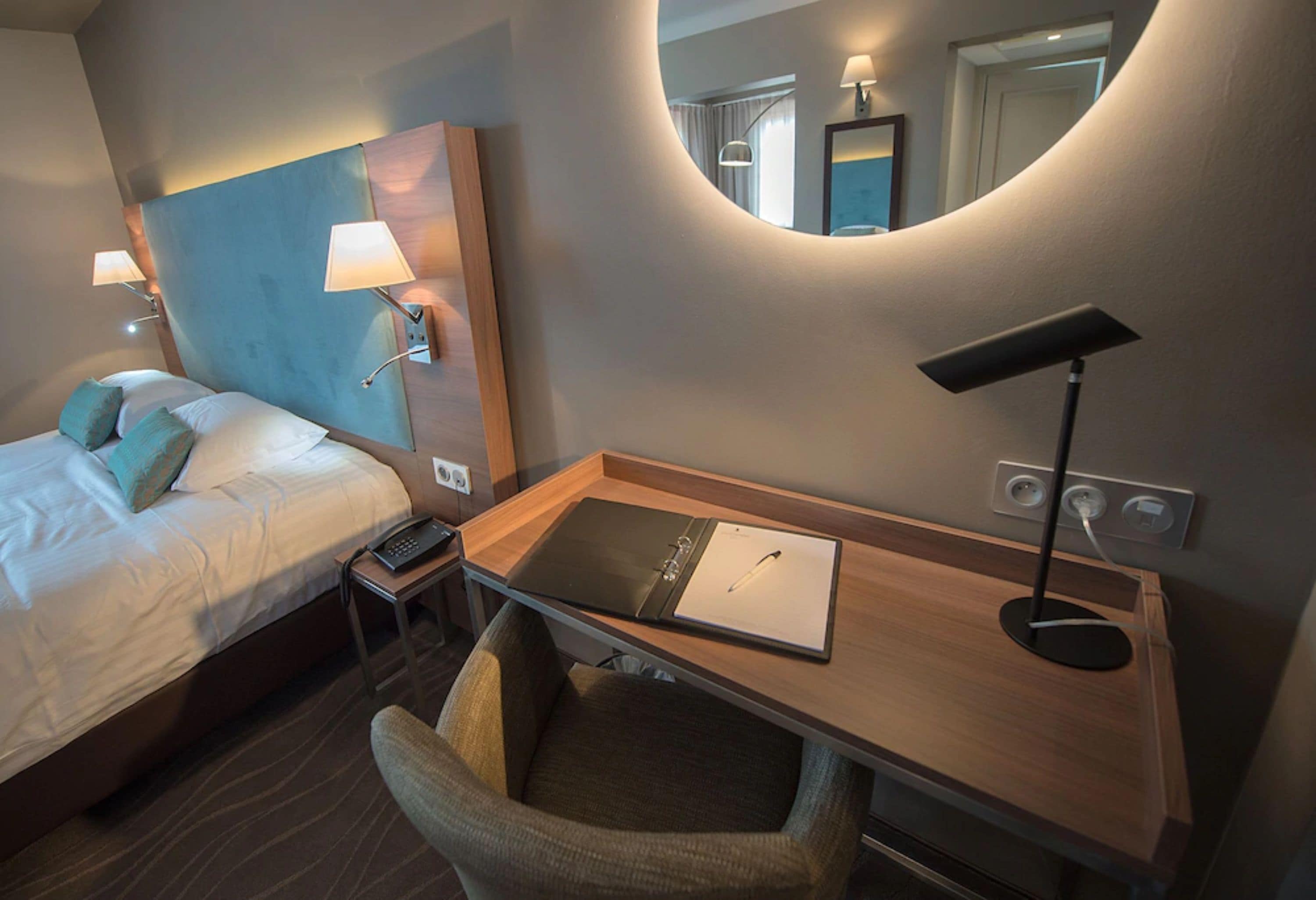 A hotel room with a bed, a desk with a lamp, a chair, a mirror, and a bedside table with a phone.