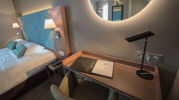 A hotel room with a bed, a desk with a lamp, a chair, a mirror, and a bedside table with a phone.