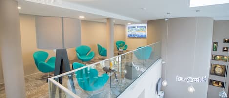 Un hall d’hôtel moderne avec des fauteuils de couleur turquoise, une rambarde en verre et un écran numérique affichant une vue panoramique de la ville.