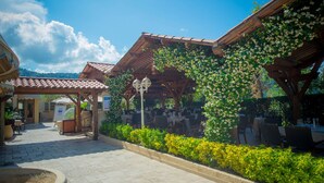 An outdoor café with tables and chairs, surrounded by greenery and a tiled roof structure. - Hôtel Lou Castelet (Carros)