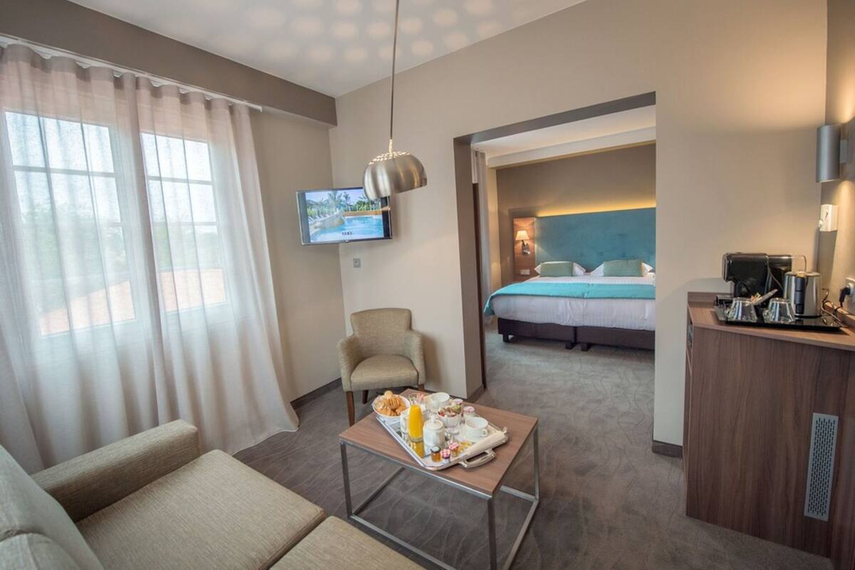 A hotel room with a bed, a TV, a dining table with breakfast, a chair, and a desk with a coffee machine.