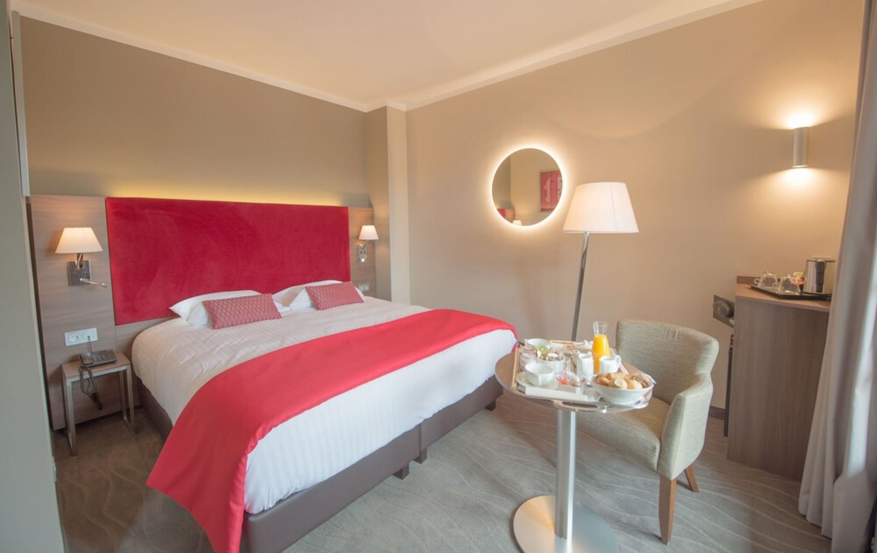 A hotel room with a large bed, a bedside table with a lamp, a small round table with breakfast items, and a chair.