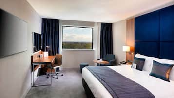 A hotel room with a large bed, a desk with a chair, a small table, a TV, and a window with curtains.