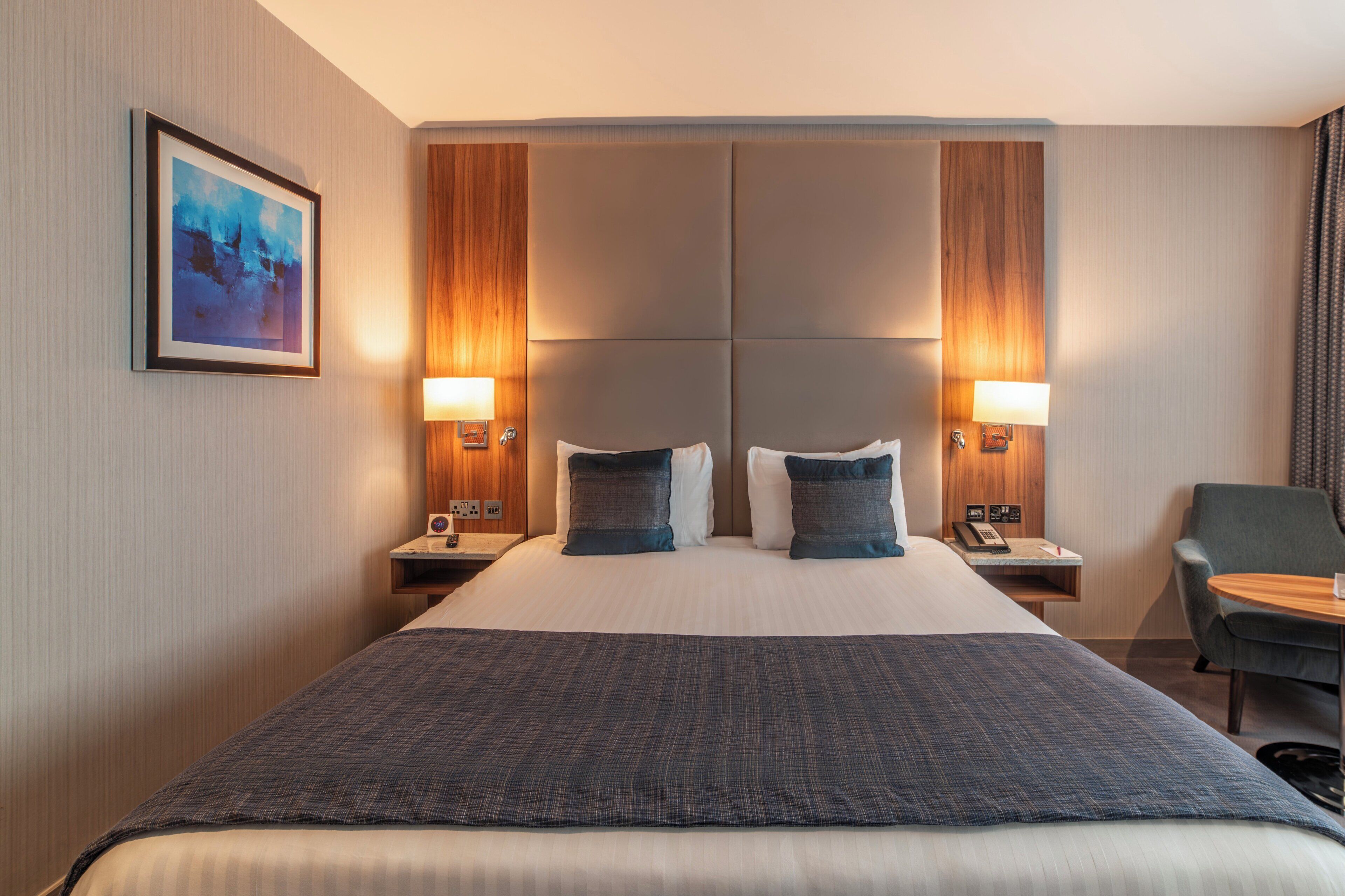 A hotel room with a large bed, two bedside tables with lamps, a chair, and a painting on the wall.