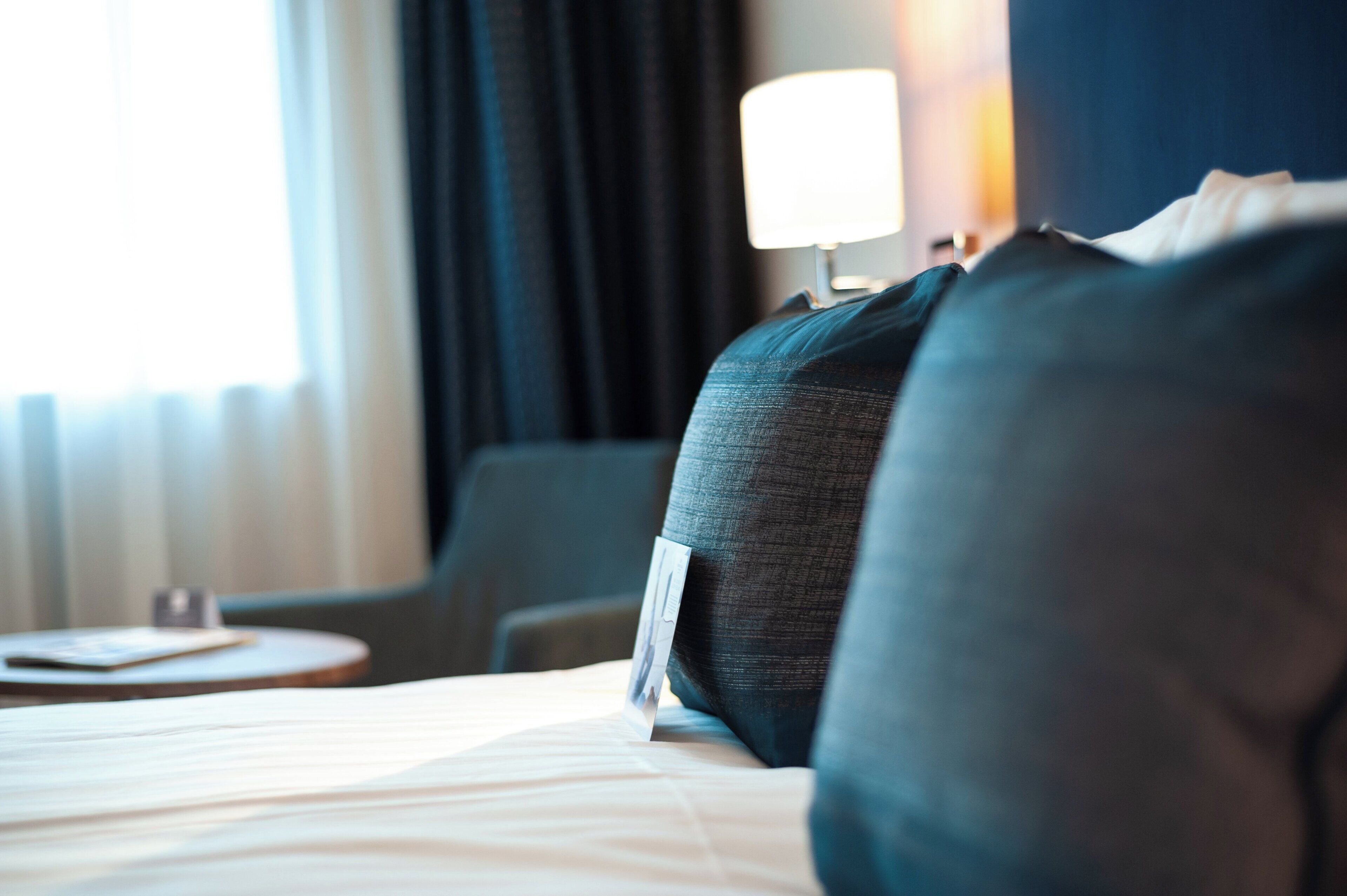 A hotel room with a bed, pillows, a bedside table, a lamp, and a window with curtains.